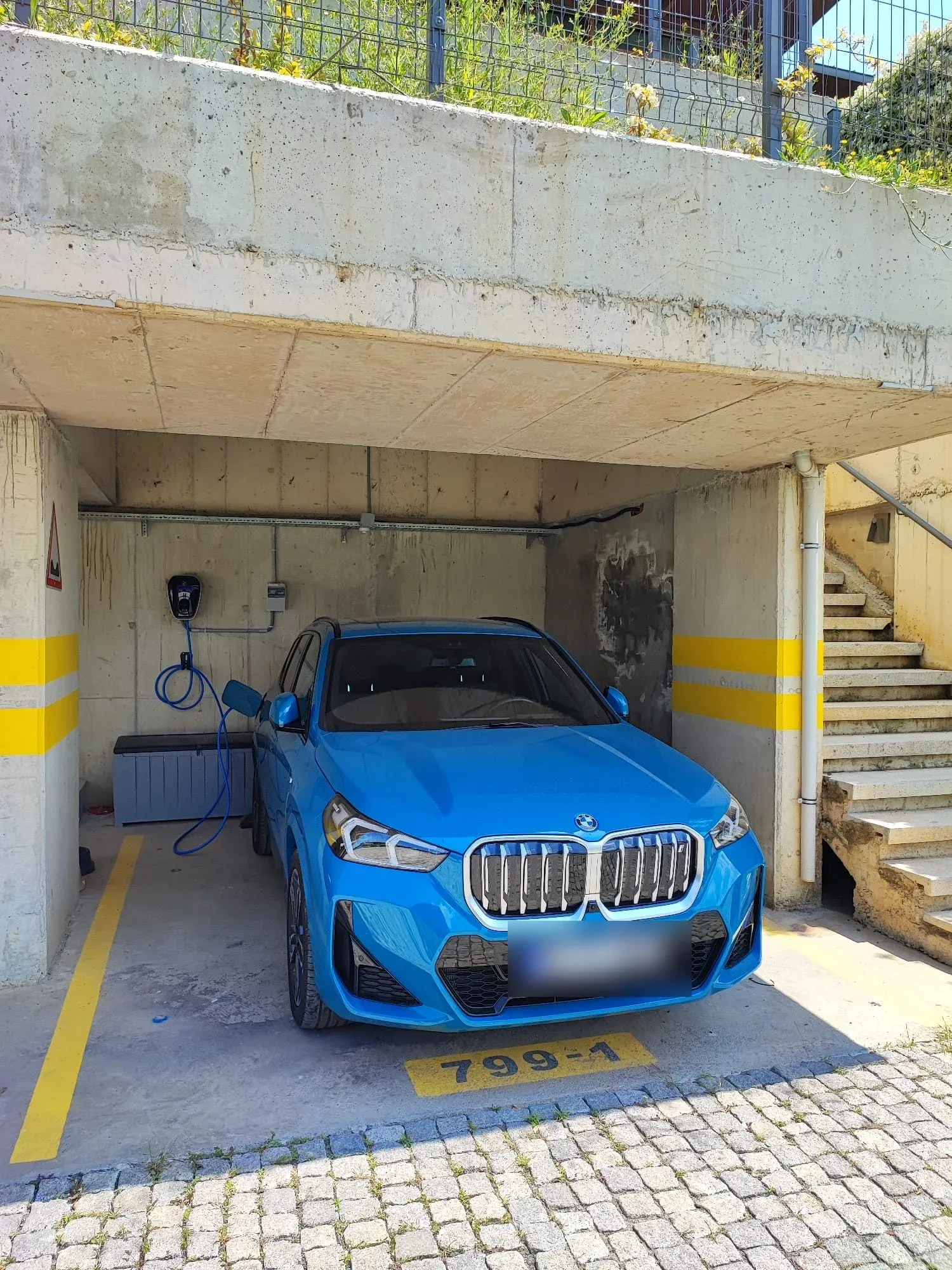 Beton bir otopark alanında parlak mavi BMW marka bir otomobil park halinde görülüyor. Aracın sol tarafındaki duvarda monte edilmiş bir şarj kutusu ve mavi kablo bulunuyor. Zemin ve sütunlarda sarı yönlendirme çizgileri, sağ tarafta beton merdivenler ve üstte tel çit yer alıyor. Aracın ön plakası bulanıklaştırılmış.